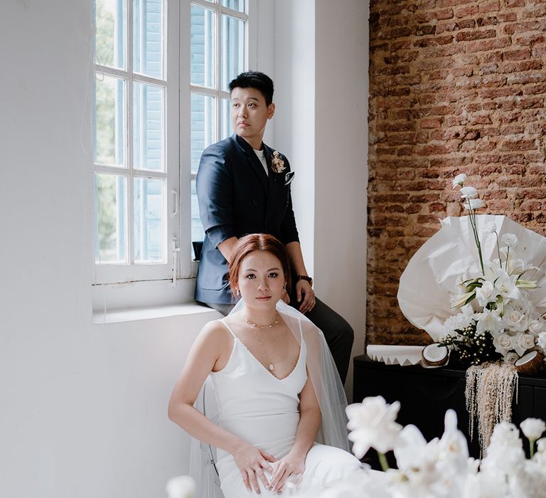 Bride in a navy suit jacket and bride in a minimalist wedding dress sitting at their all-white intimate tablescape 