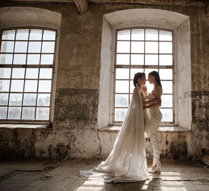 Brides embracing in front of an industrial wedding venue window in trouser separates and bridal cape 