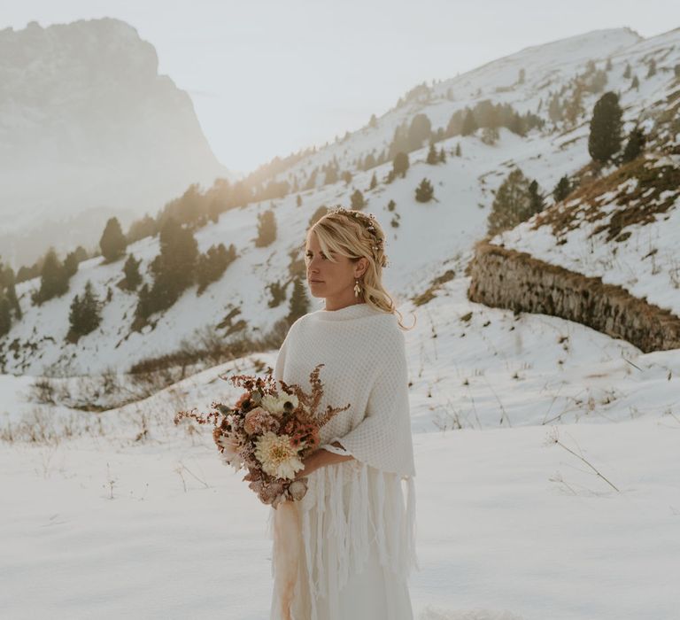 Boho bride in a cream waffle shawl holding an orange autumn wedding bouquet at snowy Dolomites wedding 