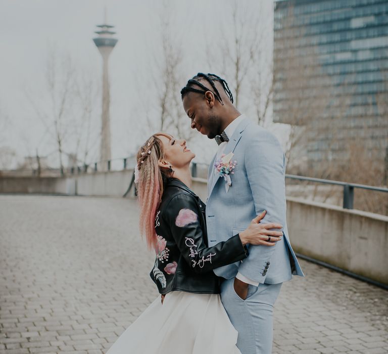 Bride with ombre pink hair wearing a customised bridal leather jacket looking up at her groom in a baby blue suit 