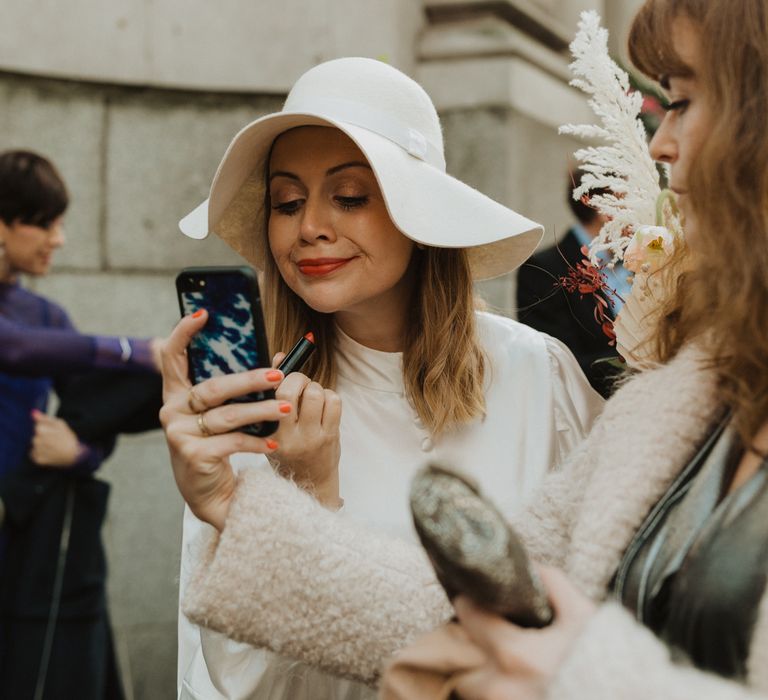 Bride reapplies her red lipstick on wedding day whilst wearing floppy hat