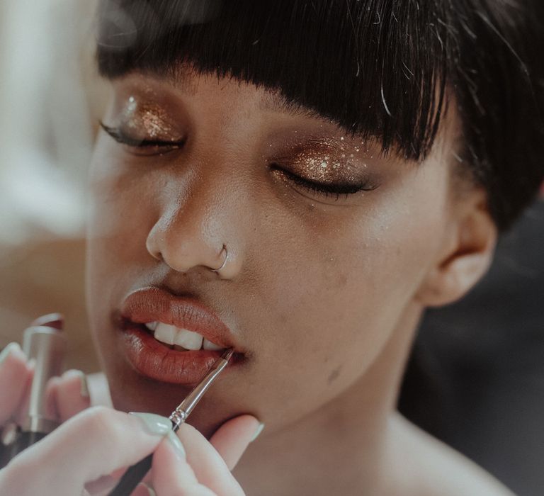 A Black bride gets her makeup done. She has golden glitter eyeshadow and is having her lipstick applied. 