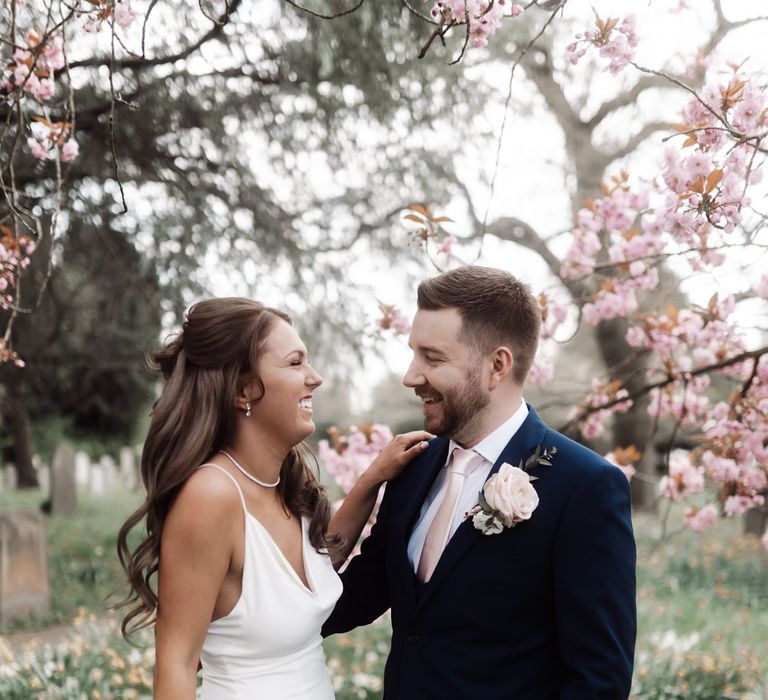 Bride and groom in church grounds surrounded by blossom and flowers, the bride is holding a bouquet of white and pink roses