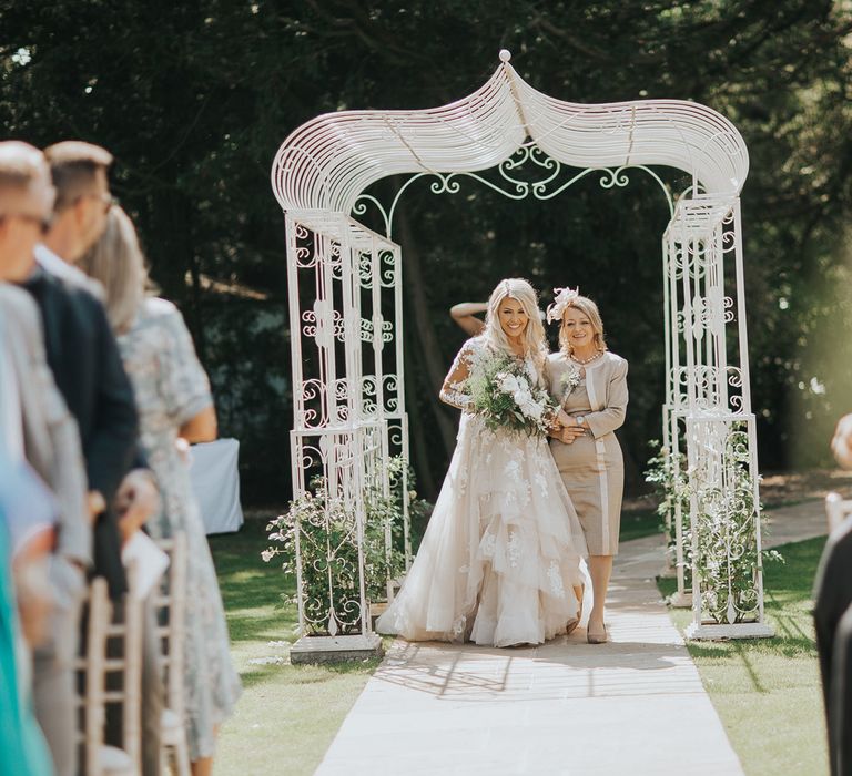 A bride wearing a long tulle layered wedding dress walks down an outdoor aisle with her mom.