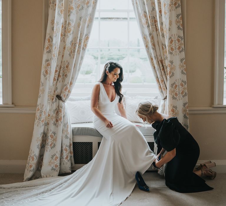 Bride with long dark curled wedding hair and white Pronovias wedding dress with train sits in bedroom at Came House Dorset with maid of honour in black one-shoulder dress helping her put on her shoes