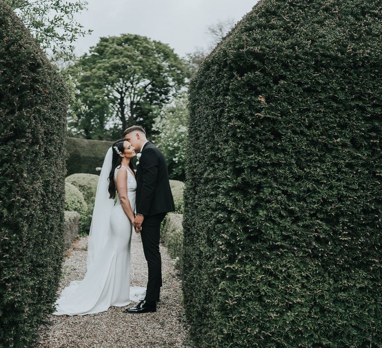 Bride in white Pronovias wedding dress with train and veil stands holding hands with groom in black tuxedo in the gardens at Came House Dorset after their wedding