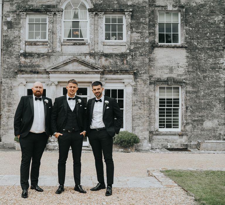 Groom and two groomsmen all in black tuxedos with white buttonholes stand in front of Came House Dorset