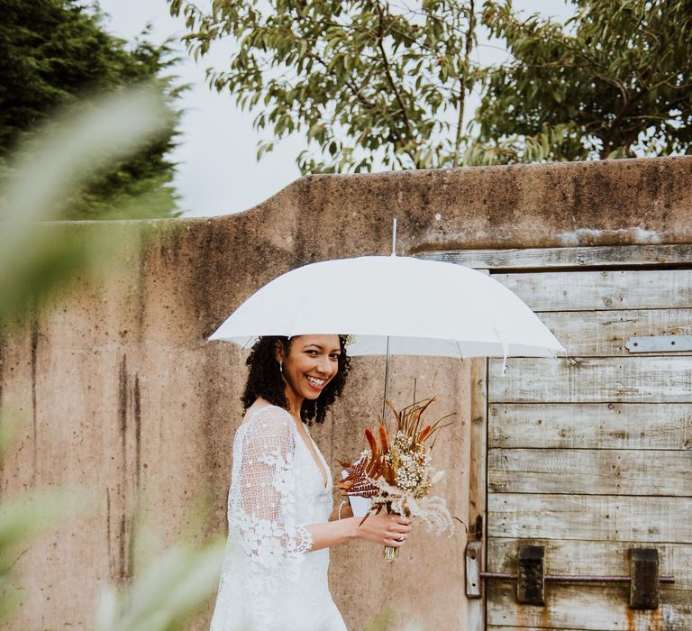 Bride stands beneath white umbrella on her wedding day whilst wearing lace gown and holding dried floral bouquet 