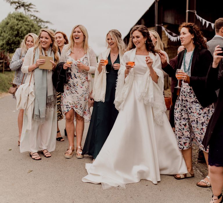 Bride in white Elbeth Gillis wedding dress and white shawl stands outside barn smiling with wedding guests as they all hold aperol spritzes at home farm wedding