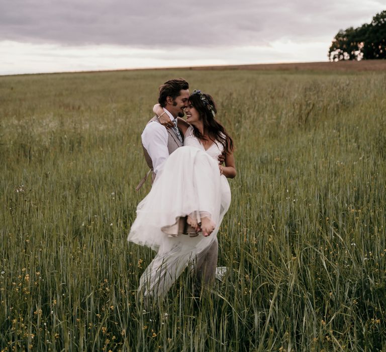 Groom carries bride through green fields on the day of their wedding