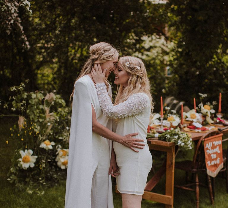 Two brides embracing in front of their outdoor tablescape with bold colours and wildflower decor 