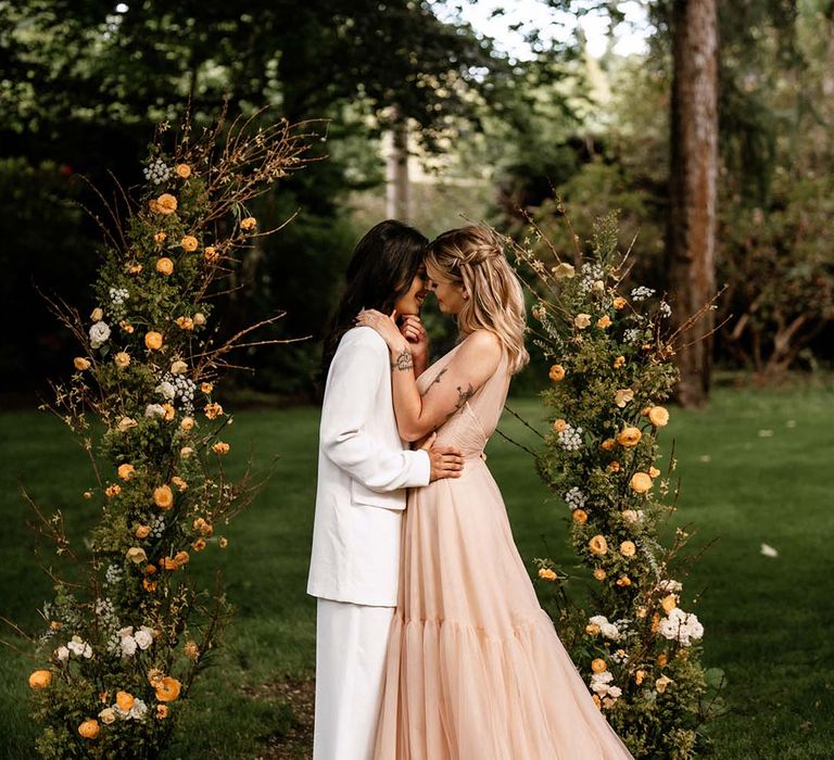 Two brides kissing in between vertical green foliage and yellow flower arrangements at their outdoor wedding ceremony 