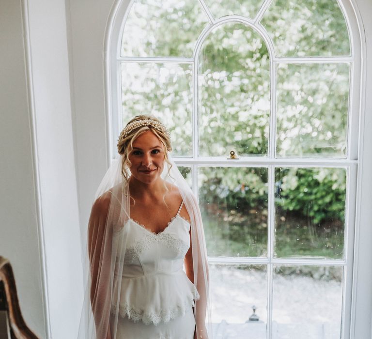 Bride stands in front of window wearing Whistles wedding gown complete with beaded headband on her wedding day