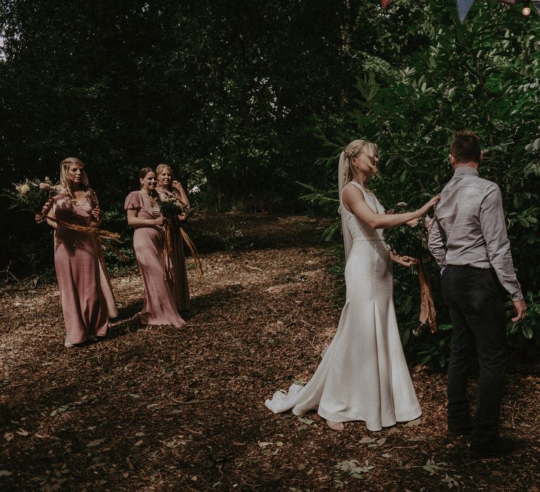 Bride taps groom on the shoulder as he turns around to see her and bridesmaids look on in woodland ceremony