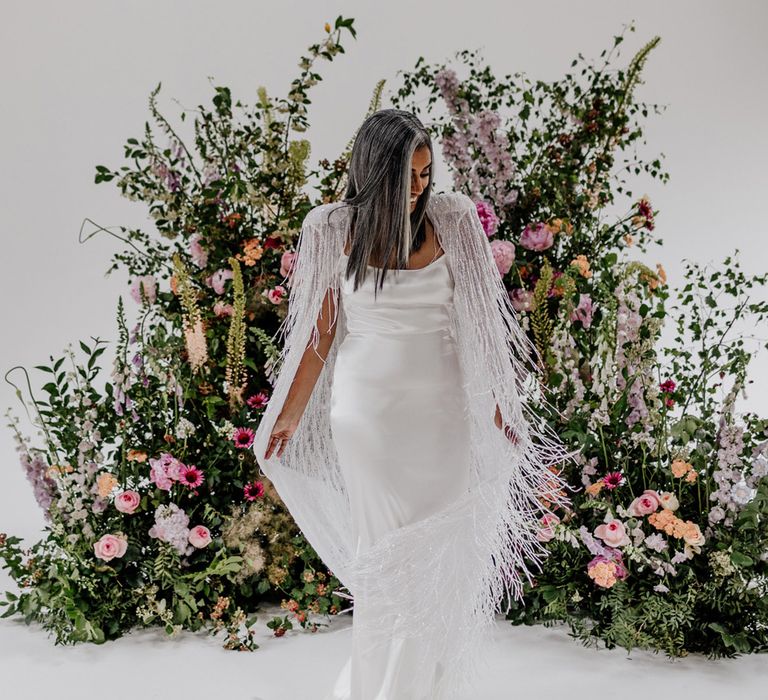 Bride in satin Halfpenny London wedding dress twirls tasselled Halfpenny London bridal cape whilst stood in front of multicoloured floral installation before wedding at Loft Studios London