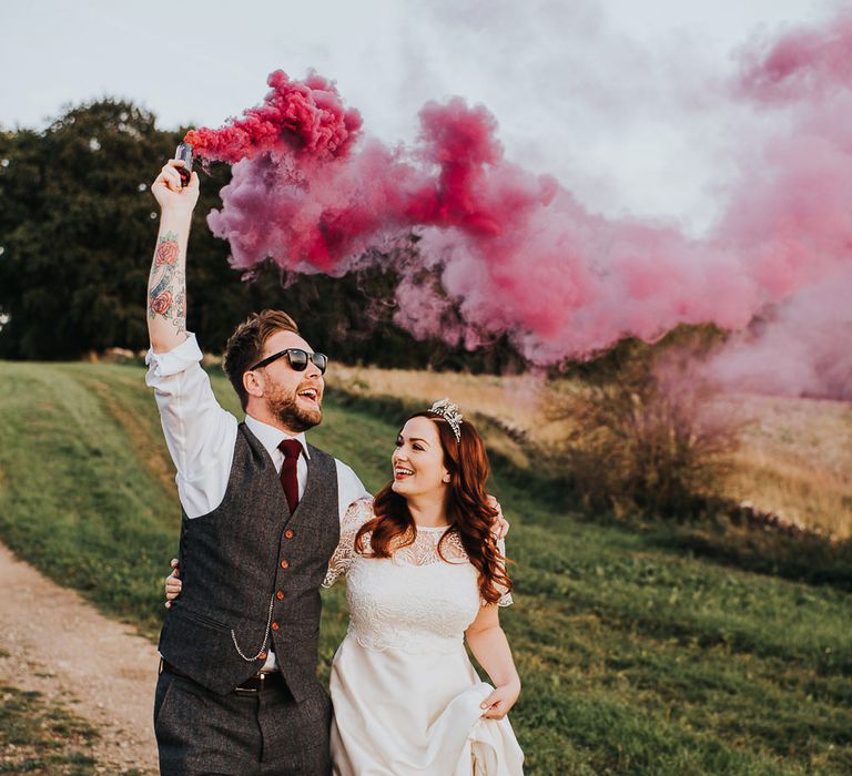 Smoke bomb couples portrait with groom in a tweed waistcoat holding a pink grenade as he walks with his bride in an A line wedding dress and bridal crown  