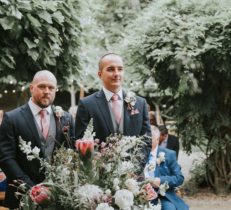 Groom in dark three piece suit with grey waistcoat and pink tie stands with best man by floral bouquet with proteas waiting for bride at Tythe Barn wedding with barn wedding flowers