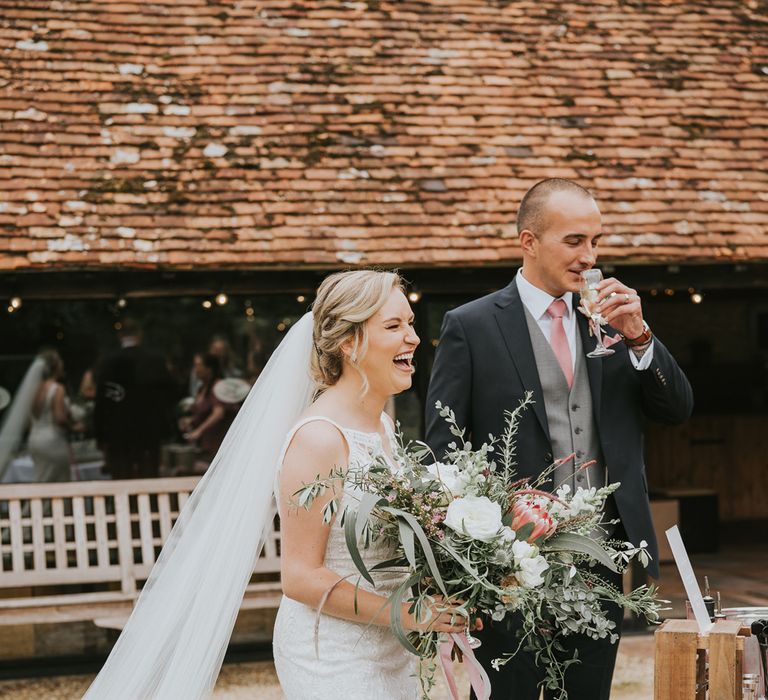 Bride in lace Justin Alexander wedding dress with veil holding pink, white and green mixed bridal bouquet stands next to groom sipping flute of sparkling wine at Tythe Barn wedding with barn wedding flowers