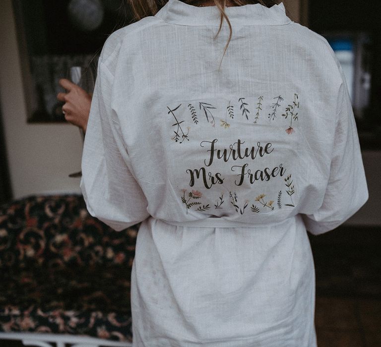 Bride in white dressing gown with writing on the back reading 'Future Mrs Fraser' surrounded by leaf illustrations before Isle of Wight wedding with macrame wedding decor