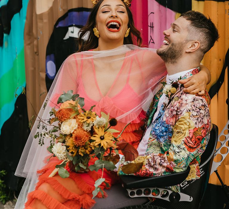 Bride in a coral wedding dress and cape veil holding a dahlia bouquet laughing with her groom in a colourful blazer 