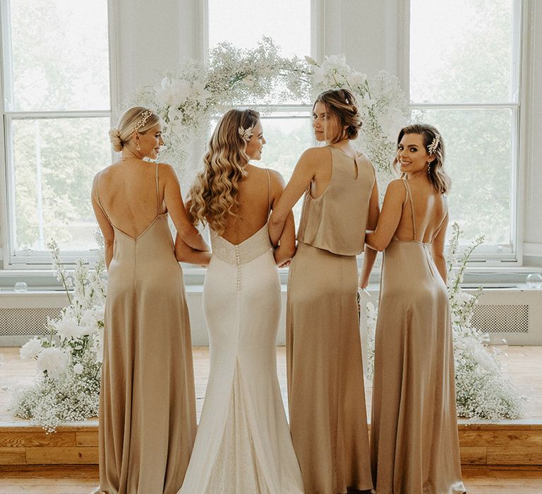 Bride in a low back wedding dress with embellishment and buttons arm in arm with her bridesmaids in champagne gold satin dresses by Constellation Ame 