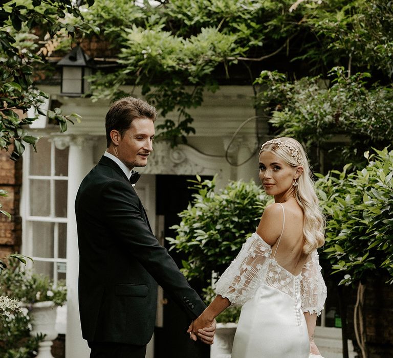 Bride in a satin slip wedding dress with lace inserts and detachable sleeves holding hands with her groom in a black suit 