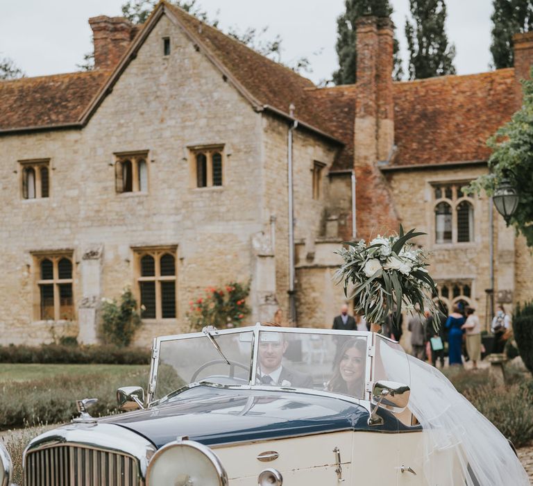 Bride in Pronovias wedding dress, Jimmy Choo wedding shoes and veil holds up white and green bridal bouquet as she sits in vintage car with groom at Notley Abbey wedding