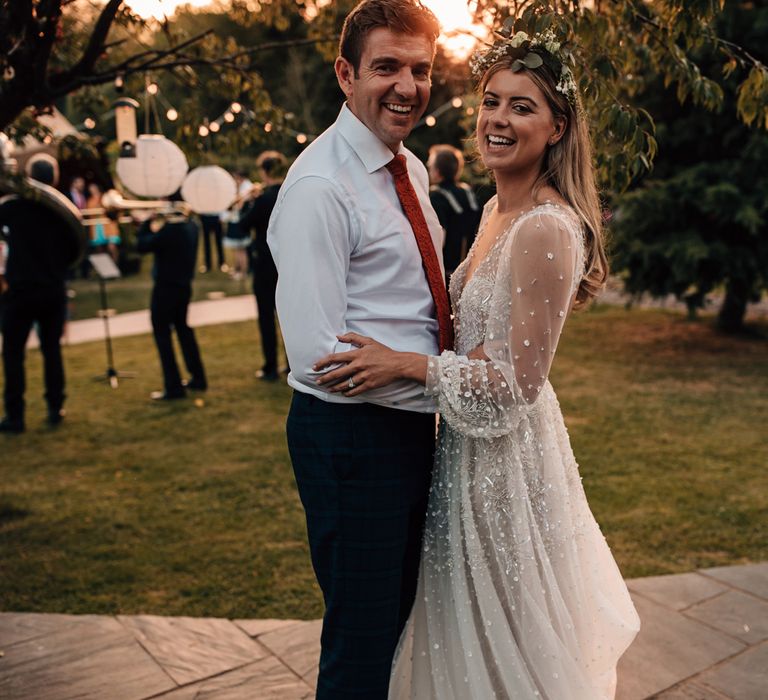 Bride in mesh long sleeve wedding dress and boho flower crown smiles whilst dancing with groom in white shirt during golden hour at tipi wedding 