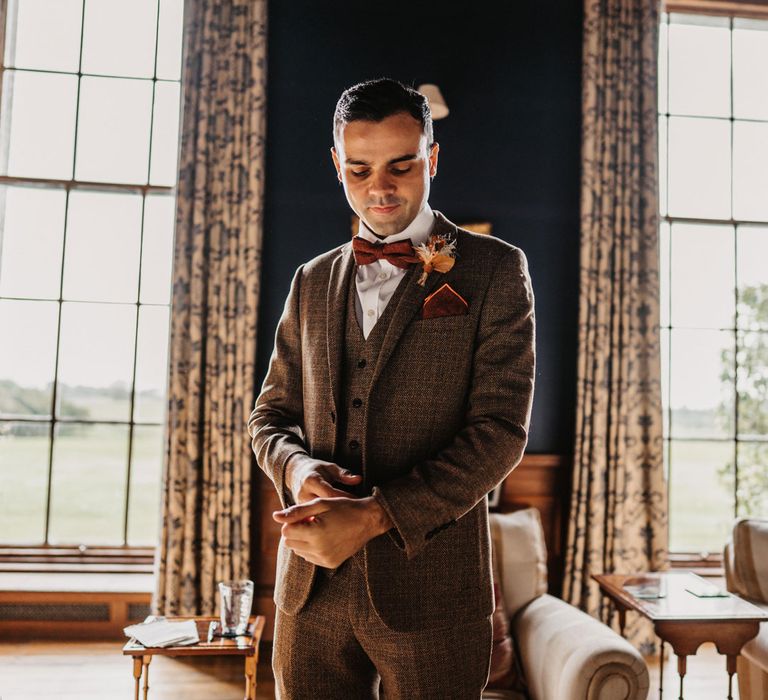 Groom in brown three piece tweed suit with orange bow tie and pocket square adjusts cuff as he stands inside room at Kinmount House before wedding