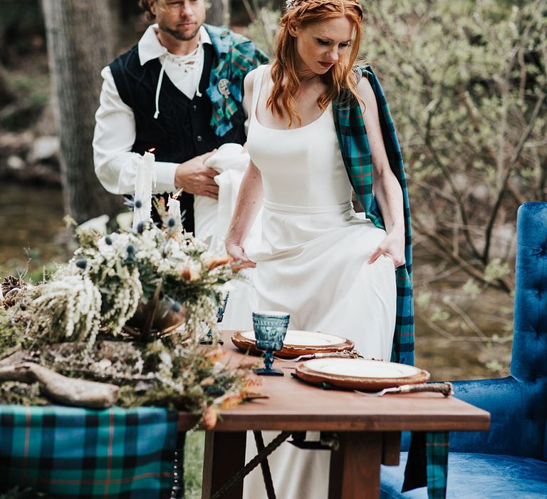 Groom dresses in traditional highland wear helping his bride in a taffeta wedding dress sit down at their sweetheart table 