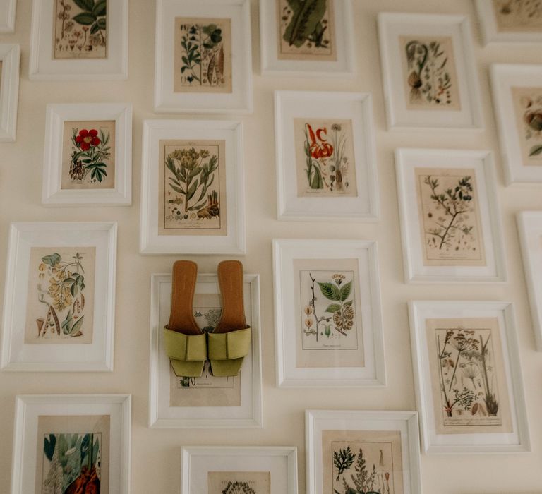 Green single strap bridal shoes hang on white picture frame next to array of framed wildflower images