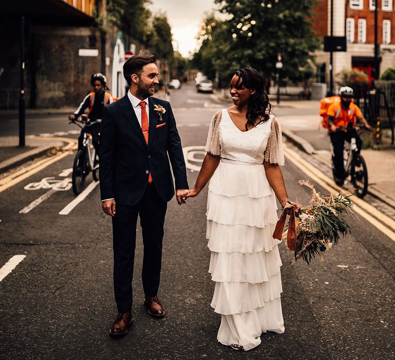 Black bride in a layered skirt and pearl top holding hands with her groom in a navy suit on the streets of London during golden hour 