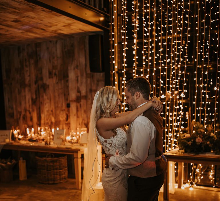 Bride in lace wedding dress with train and veil stands with groom in white shirt and waistcoat in front of fairy light curtain at rustic wedding