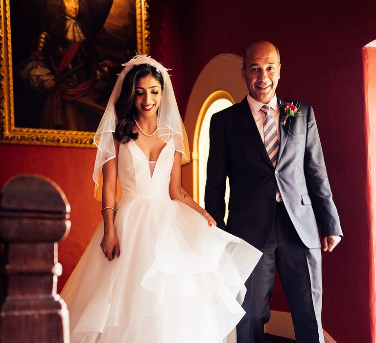 Bride in layered tulle wedding dress, pearl necklace and veil walks down staircase with man in dark suit, striped tie and buttonhole before wedding ceremony