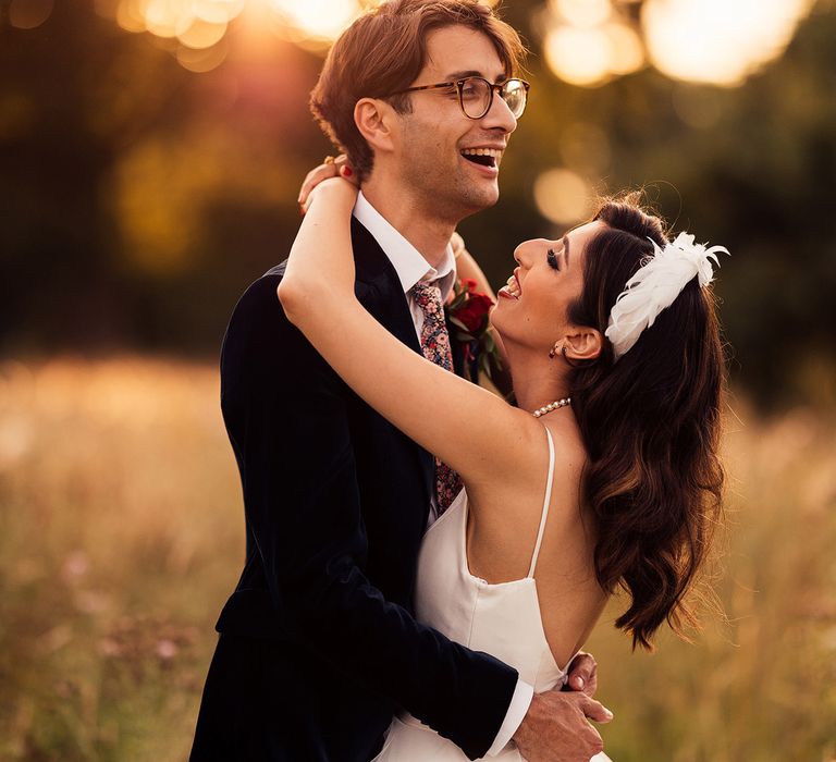 Bride in feathered bridal headband stands with arms around groom in dark blue suite, floral tie and buttonhole during golden hour