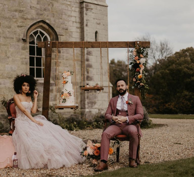 Chiddingstone Castle wedding inspiration with wooden frame cake display stand and the bride and groom sitting on ornate chairs in a pink suit and bridal separates 