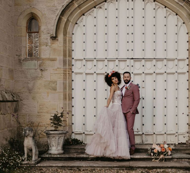 Black bride with afro hair in a pink tulle skirt and corset top embracing her groom in a pink three-piece suit at Chiddingstone Castle 