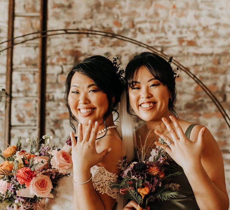 Bride in lace bardot wedding dress holding mixed floral bouquet stands with bridesmaid in sage green satin bridesmaid dress both showing off their rings