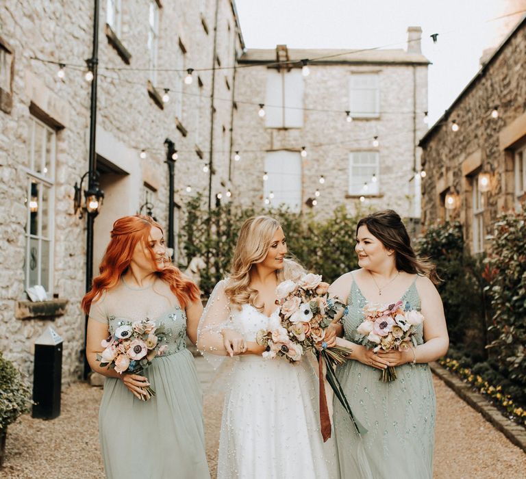 Bride in v neck wedding dress encrusted with pearls walks with bridesmaids in sage green bridesmaid dresses all holding mixed pastel wedding bouquets