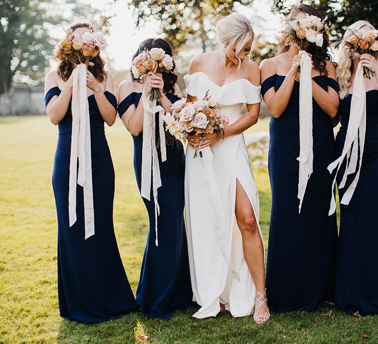 Bride in front slit wedding dress standing with bridesmaids in navy blue dresses holding up bouquets