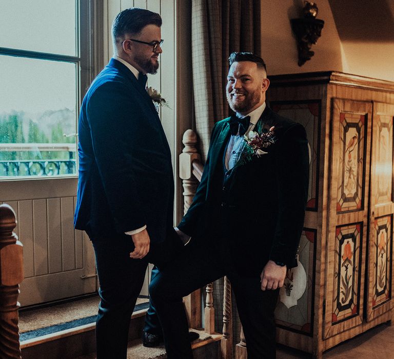 Grooms in colourful velvet suit jackets and matching black bow ties with large buttonholes pose on stairs together