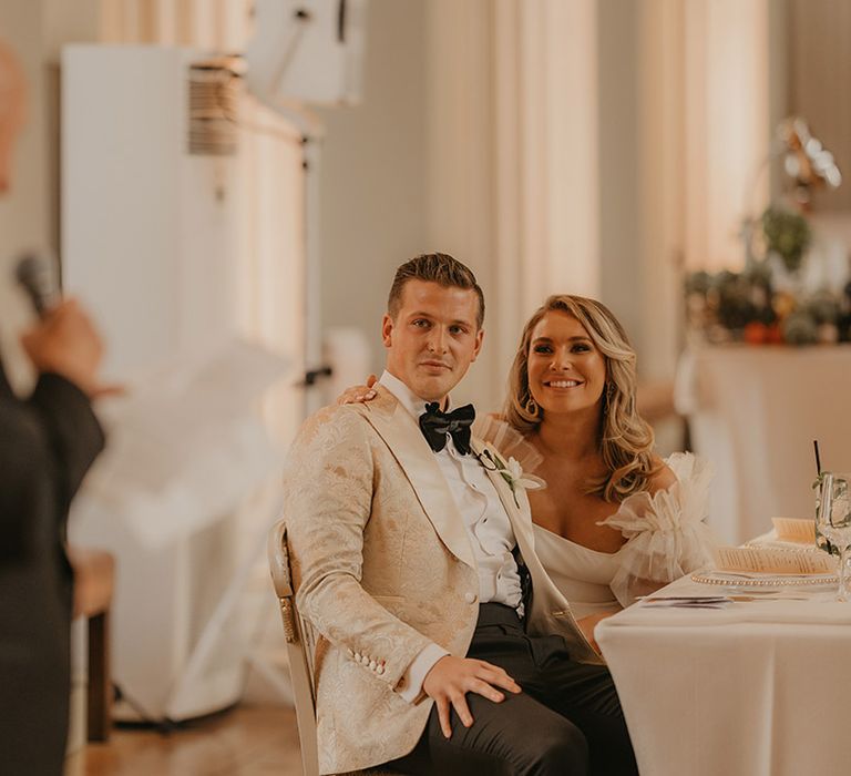 Bride and groom sit together at wedding reception as they listen to the groom's grandfather's speech 