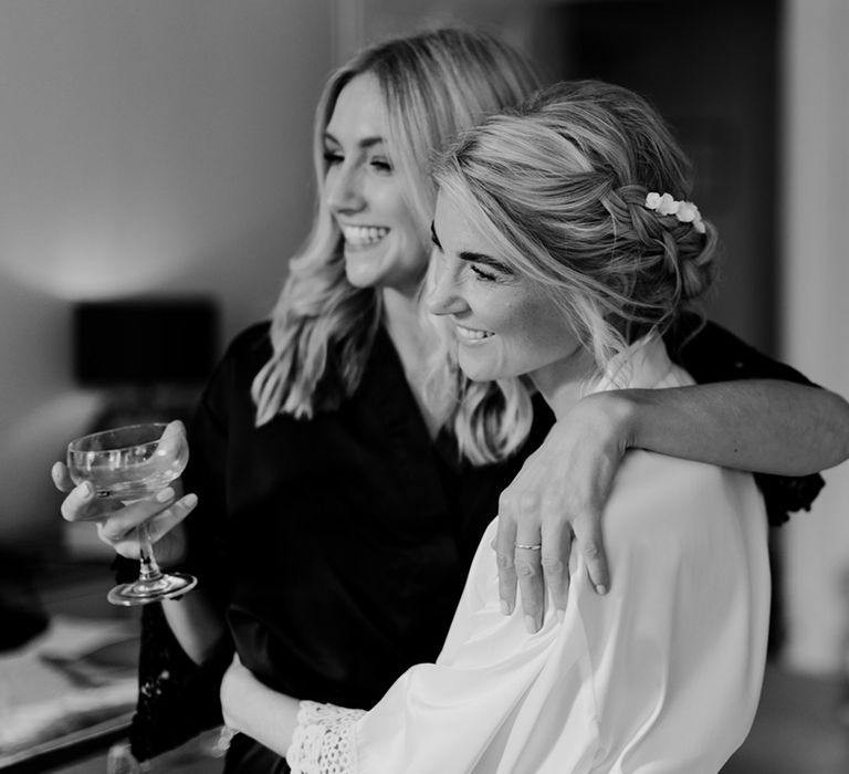 Bride in white robe hugs bridesmaid in dark robe holding glasses of champagne