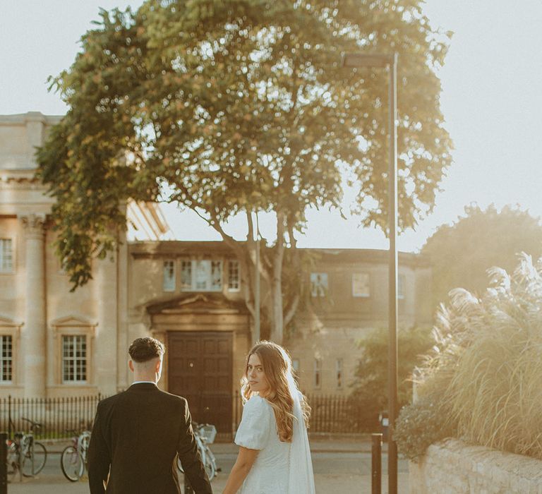 Bride looks back at the camera as she walks with groom around Oxford wearing Story of My Dress gown 