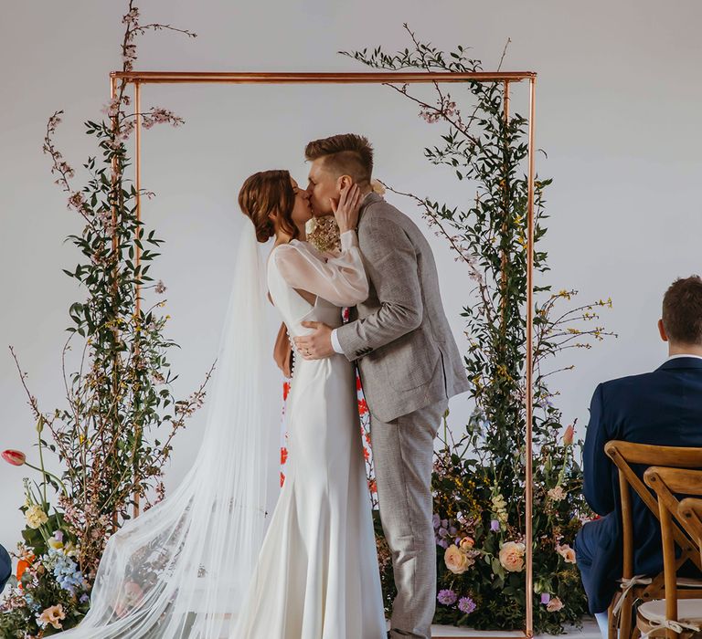 Bride in Suzanne Neville dress and long veil shares first kiss with groom wearing Reiss suit as a married couple 