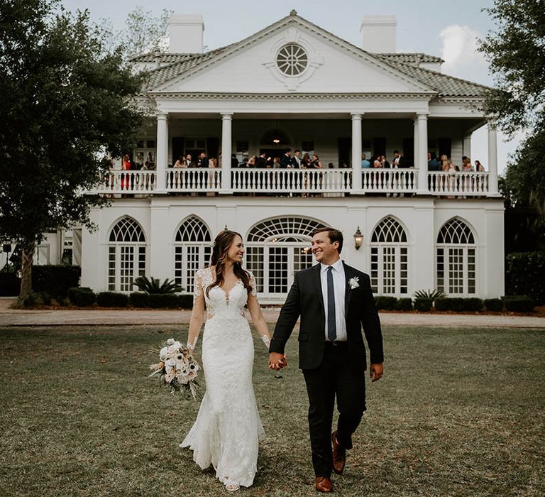 Bride in floral lace wedding dress and groom walk holding hands in front of the wedding venue with guests standing on balcony 