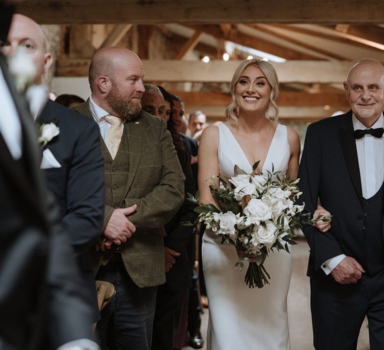 Father of the bride in black tie walks the bride down the aisle as she smiles at the groom 