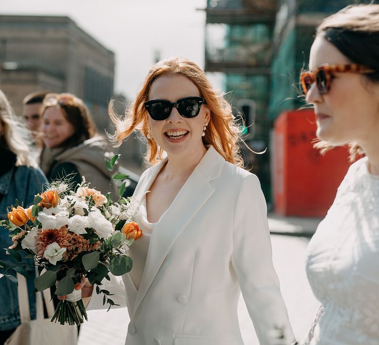 Bride looks toward camera wearing sunglasses whilst holding bright floral bouquet 