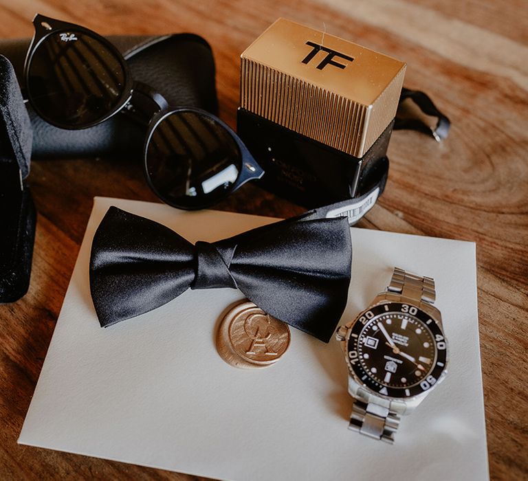 Groom's accessories with a bow tie, watch, cologne, wedding bands and gold wax seal wedding stationery