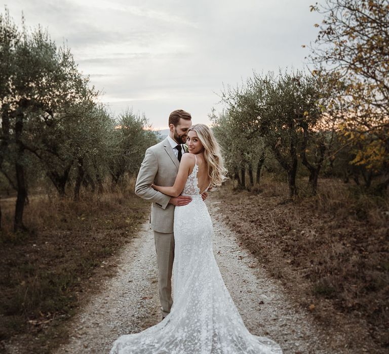 Bride & groom stand outdoors in Tuscan countryside as bridal wears Emmy Mae Bridal wedding dress complete with fanned out train and low back